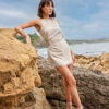 This dress is a sleeveless, natural beige mini-dress made from a lightweight fabric. It features a gathered, ruched detail at the side with an open crochet accent, adding a unique and stylish texture. The bottom hem has a delicate fringe trim, giving the dress a relaxed, bohemian vibe. The simple round neckline and fitted silhouette highlight its minimalistic yet elegant design, perfect for casual or semi-formal summer wear.