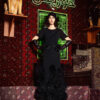 A woman in a black dress and gloves stands in a room with ornate rugs and a neon sign reading "KHOJURIS.
